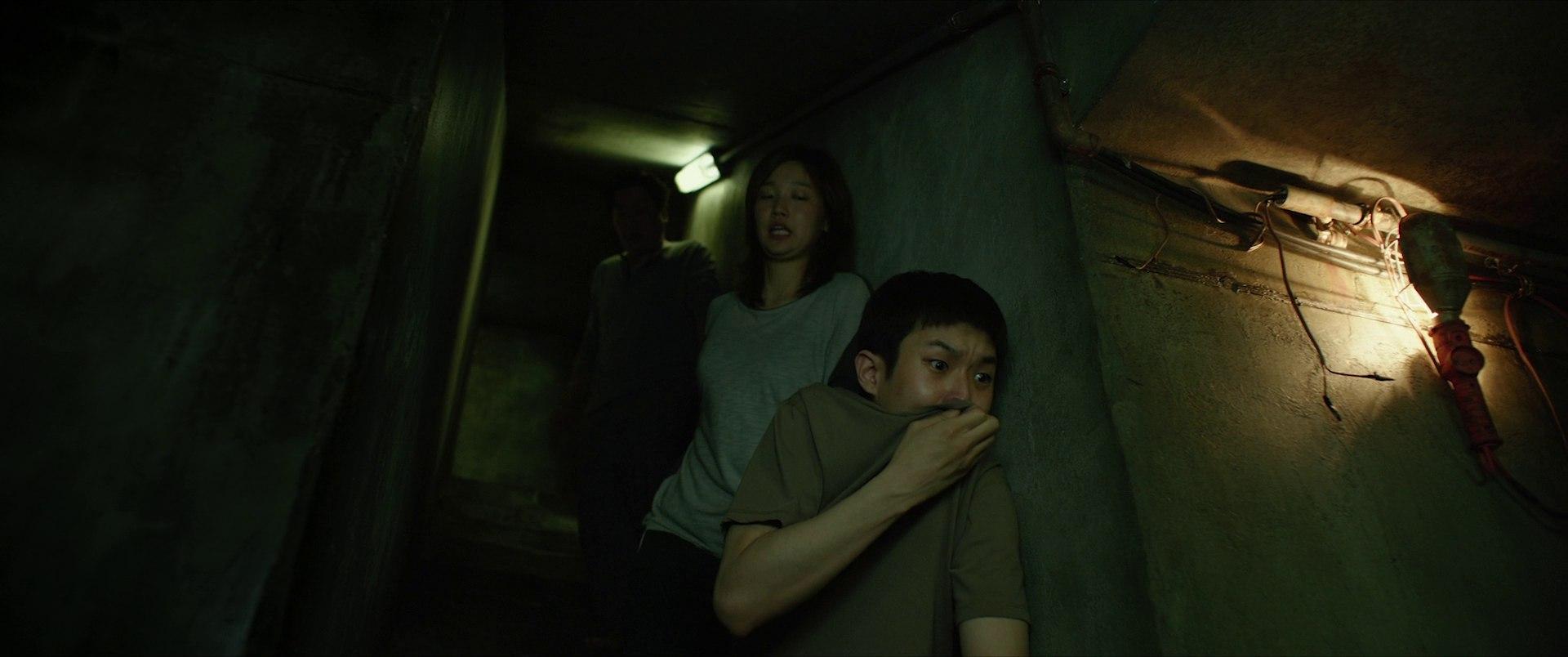 Two people pressed against the wall in the dark basement corridor, terrified